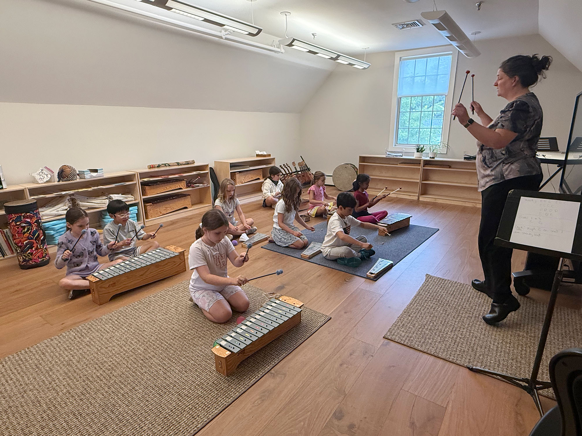 A teacher leads a group of children playing xylophones in a bright classroom with wooden shelves and flooring.