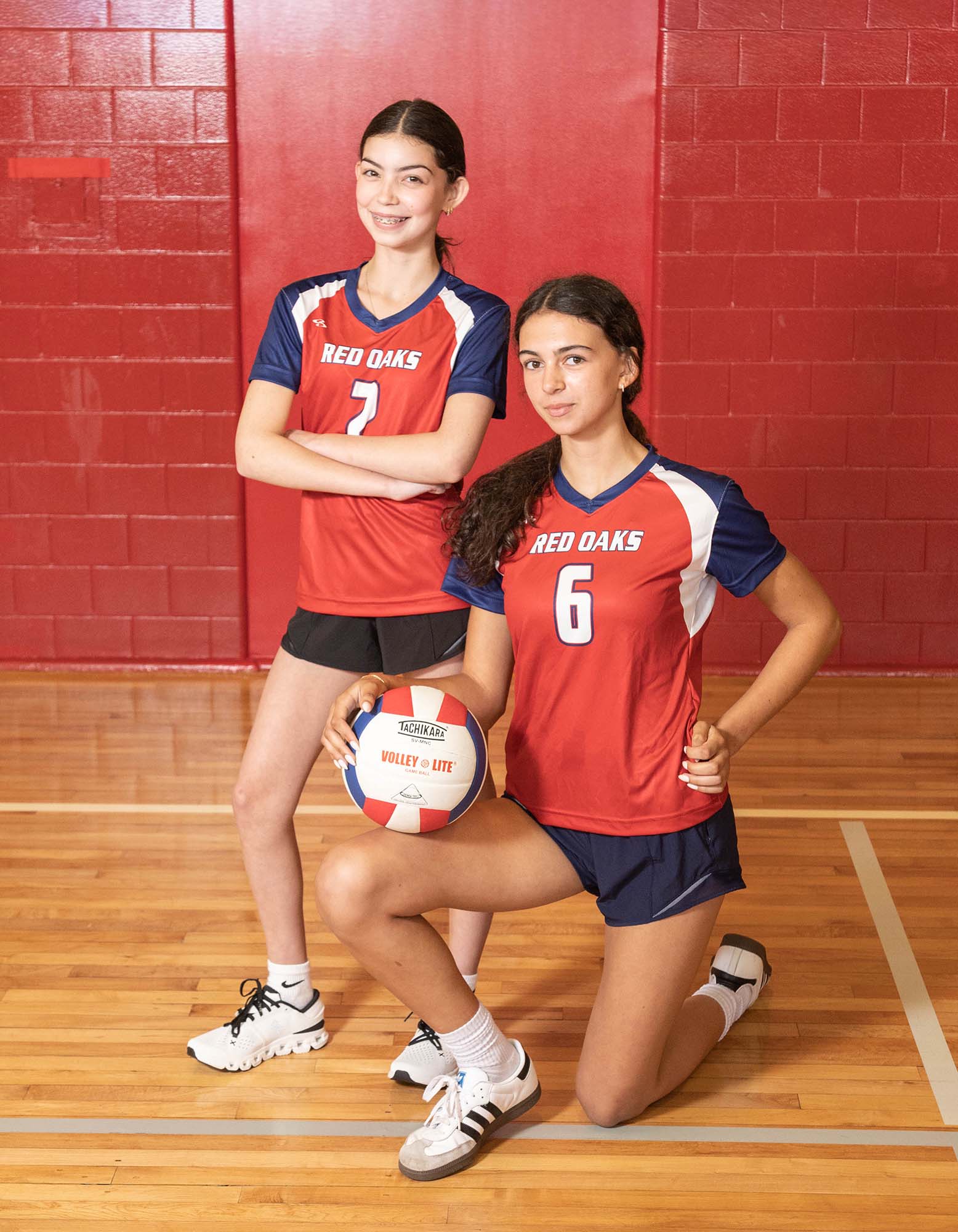 Two female volleyball players in red and blue uniforms pose with a ball on a wooden gym floor against a red brick wall.