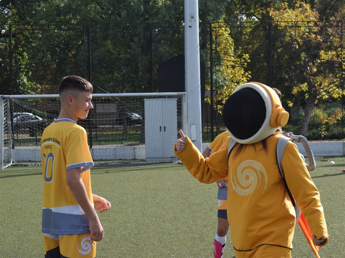 A boy in a yellow sports jersey stands facing a person in a yellow astronaut costume on a green sports field with trees and a fence in the background.