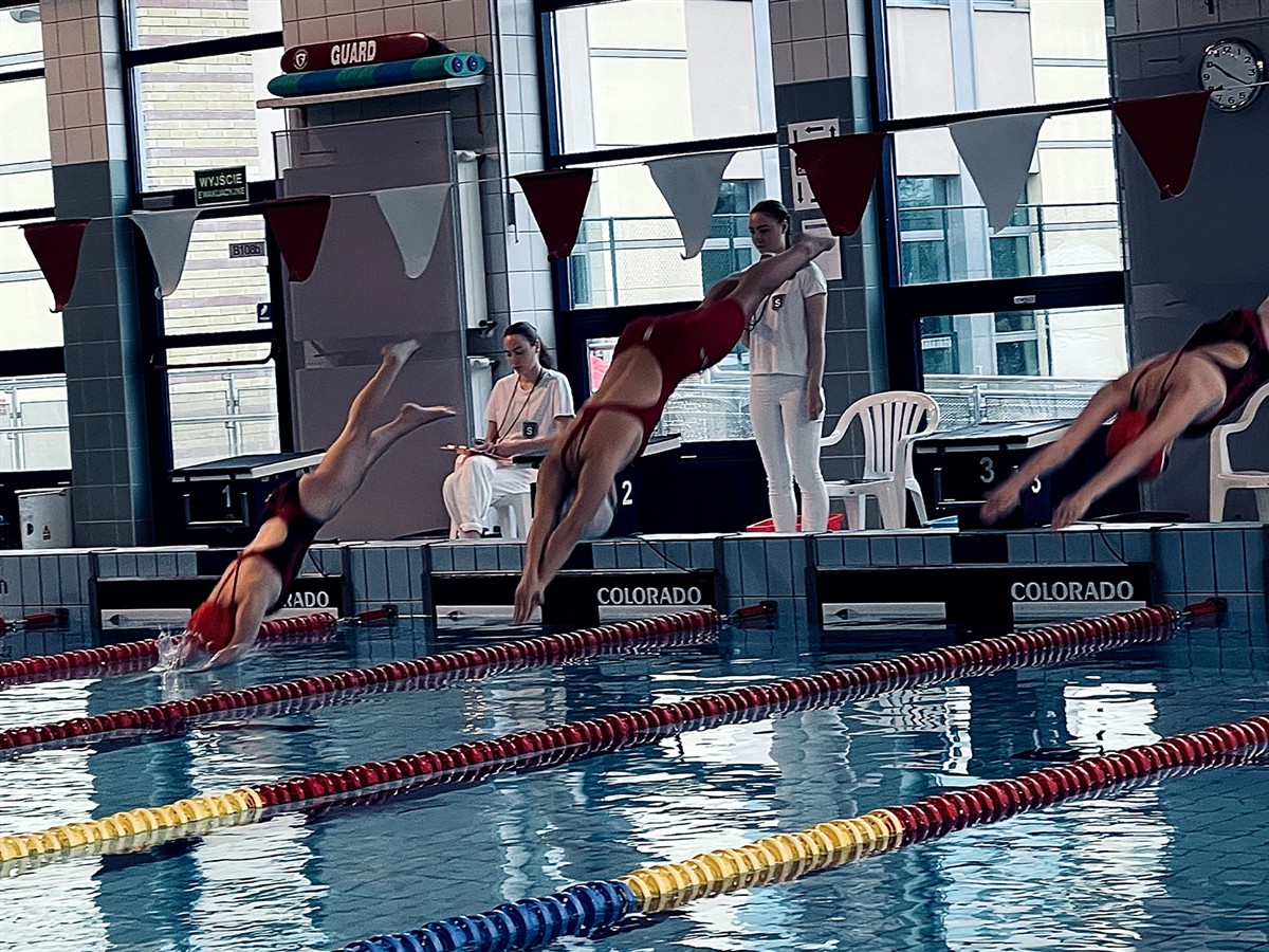 Three divers in red swimsuits leap into a swimming pool with lane markers in the foreground, while officials stand by the pool deck in a tiled indoor facility.