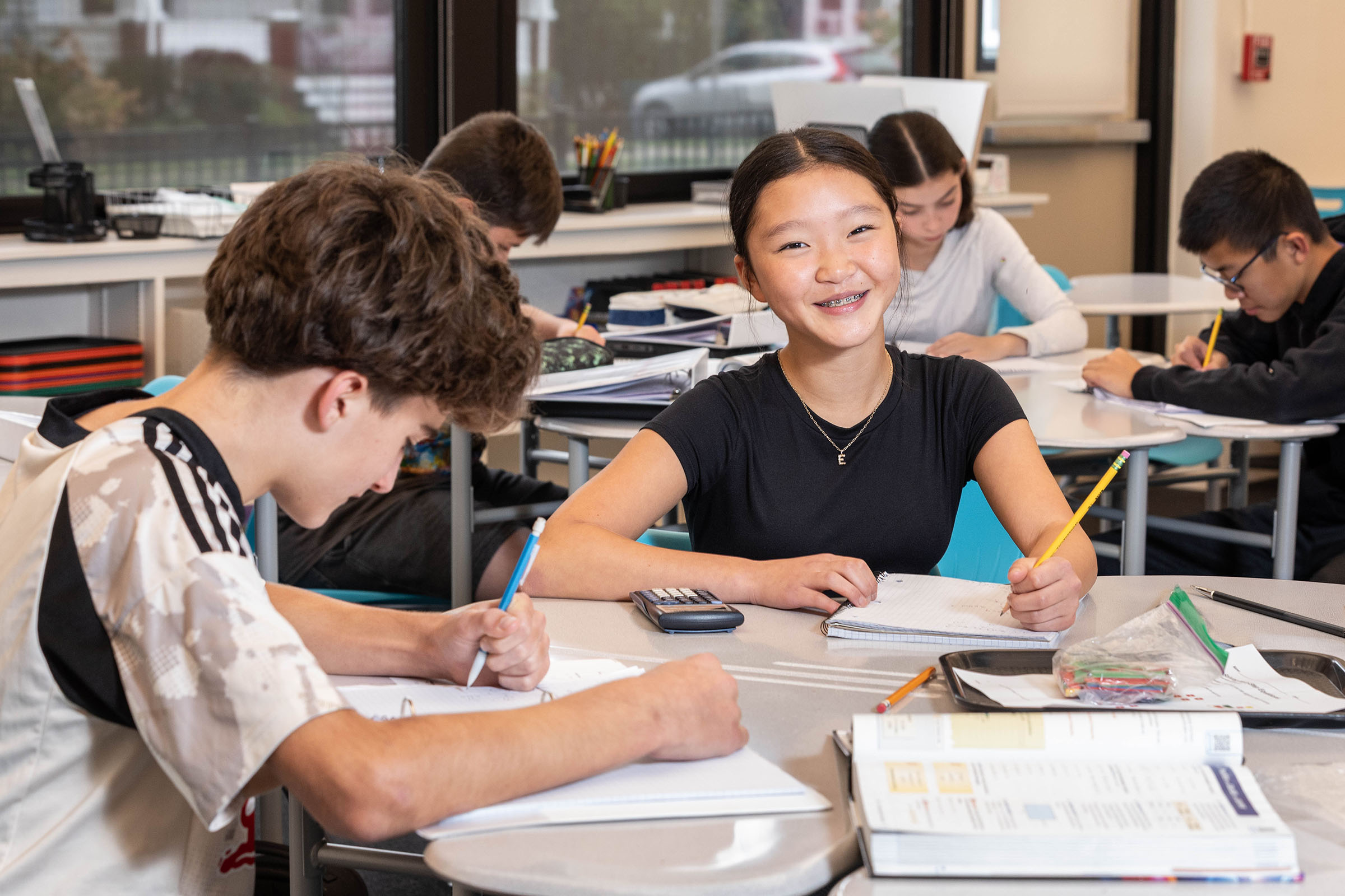 A smiling teenage girl sits at a classroom desk with a calculator and notebook, surrounded by other students studying in a bright classroom.