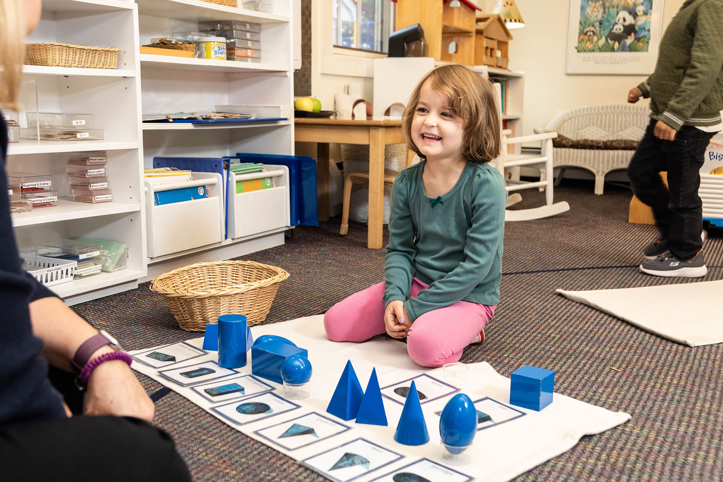 A smiling young girl sits playing with blue Montessori shapes in a classroom filled with shelves and manipulatives.
