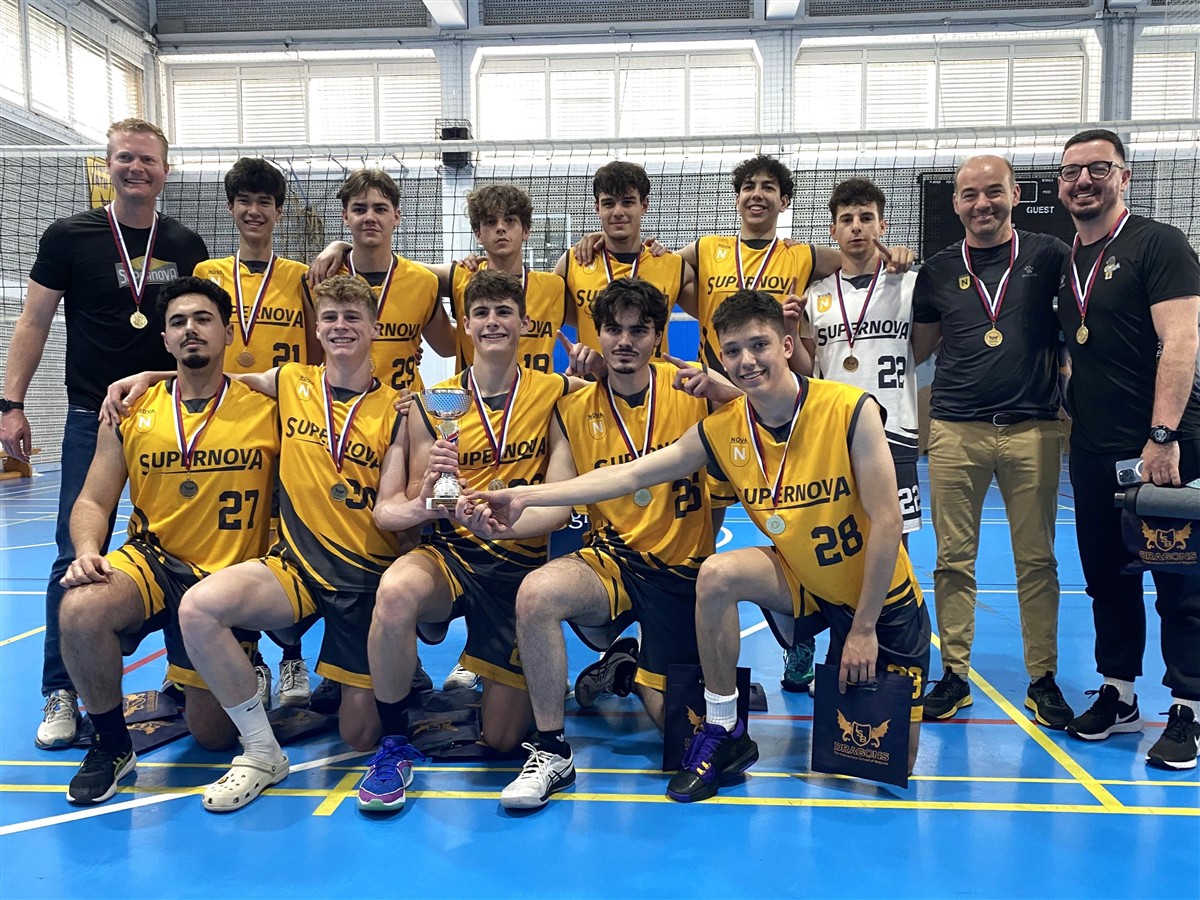 A basketball team in yellow and black uniforms poses with a trophy and medals in the foreground of an indoor sports court.