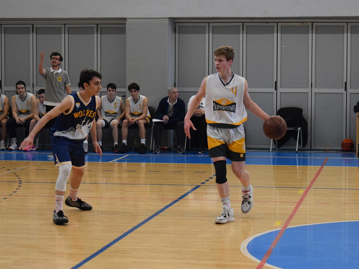 Two basketball players compete on a court while teammates and spectators watch from a bench in the background.