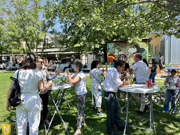 People gather around high-top tables on a sunny lawn with green trees and a colorful mural in the background.