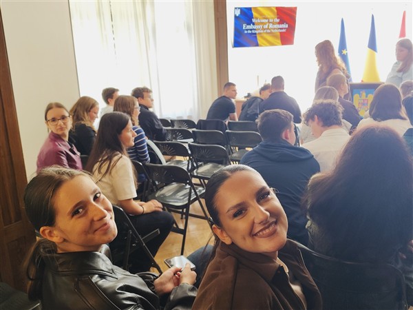 Two smiling women pose for a photo in the foreground while a group of people sit in chairs facing a stage with flags in the background.