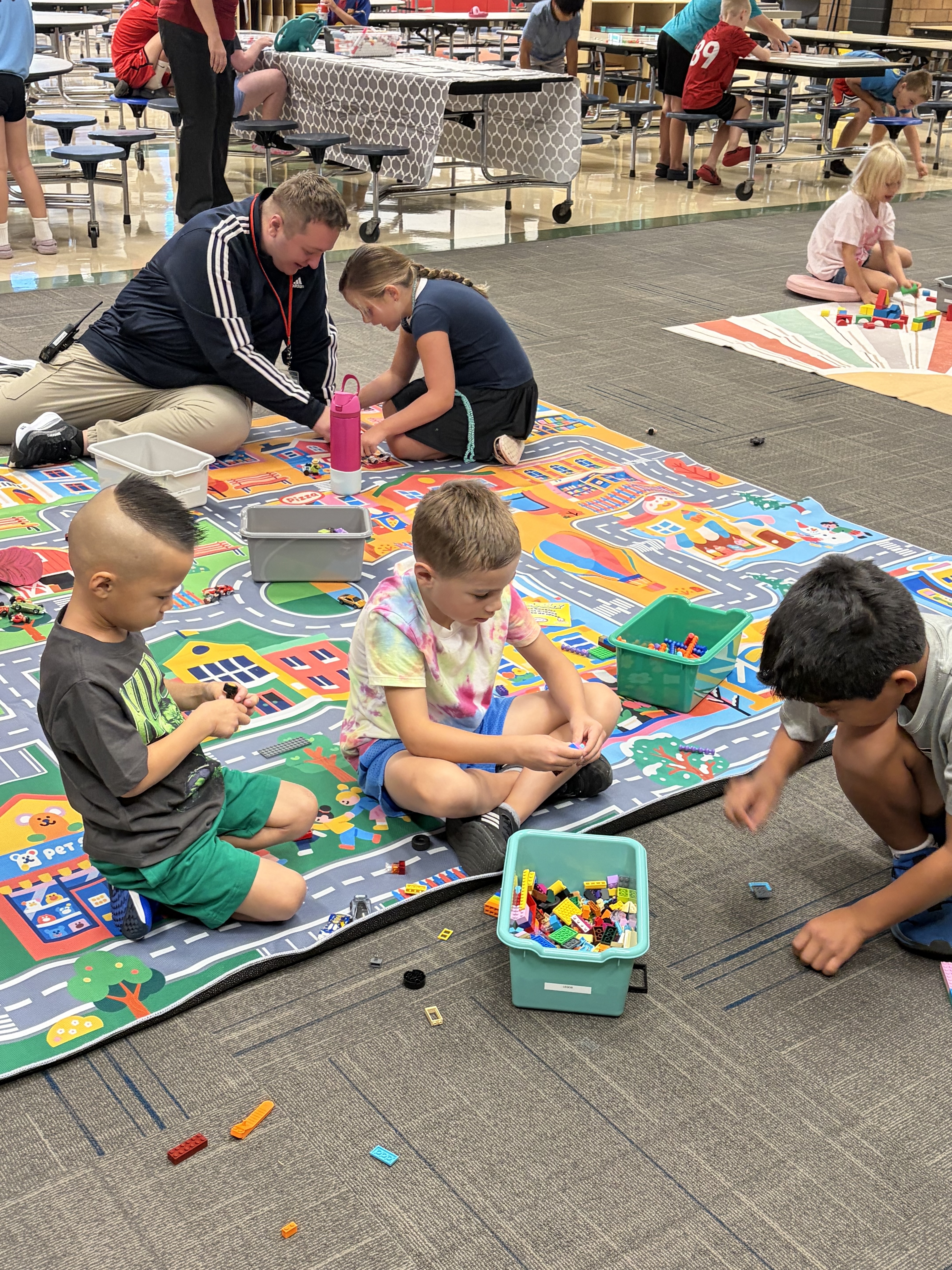 Children play with colorful building blocks on a patterned rug in the foreground, while other students and adults gather around tables in the background of a large room.