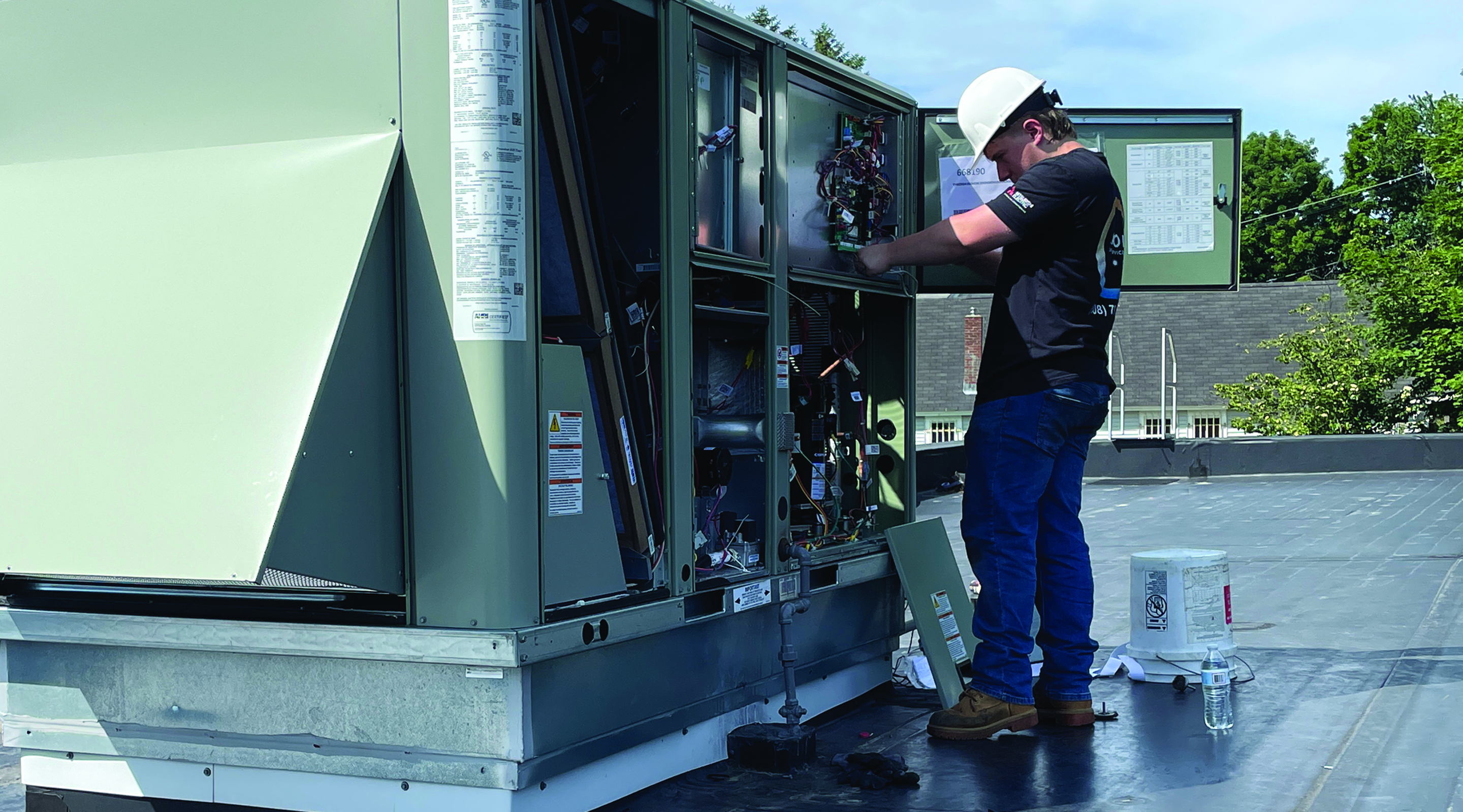 A technician in a hard hat works on a large HVAC unit on a rooftop under a clear blue sky.