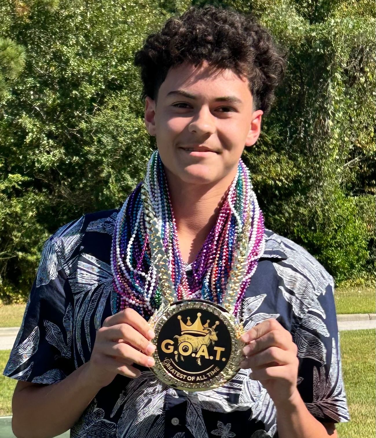 A smiling young man wearing a floral shirt and multiple colorful beaded necklaces holds a large gold "G.O.A.T." medal against a background of lush green trees.