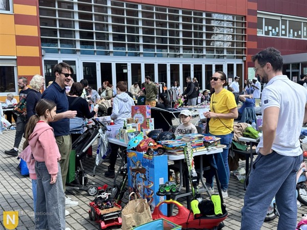 People browse a variety of toys and items at an outdoor market in front of a modern building.