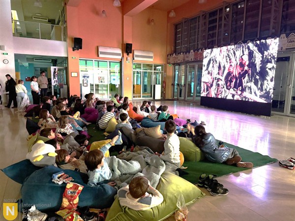 Children lounge on colorful beanbags in the foreground while watching a large screen in a spacious, modern hall.