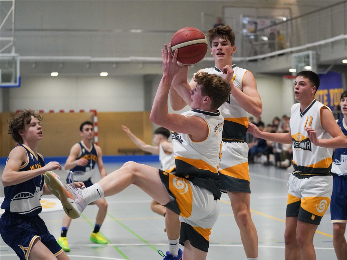 A basketball player in a white and yellow uniform leaps to shoot the ball in the foreground, surrounded by other players on an indoor court with a neutral-toned gym background.