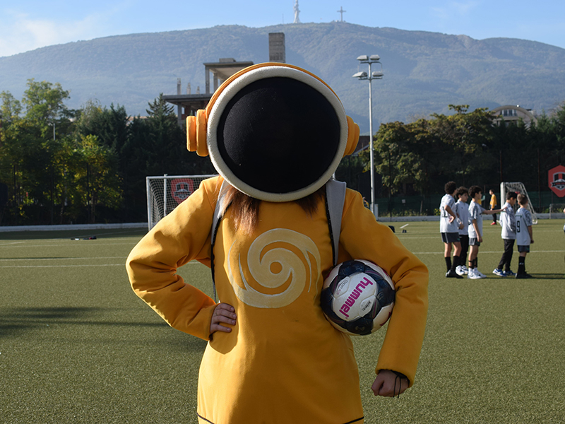 A person wearing a yellow costume with a large circular black head mask holds a soccer ball in the foreground of a green sports field with players and mountains in the background.